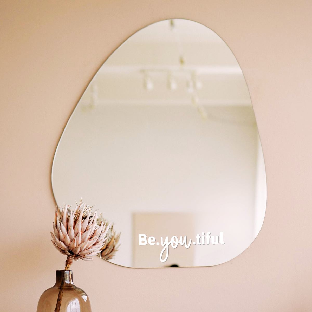 Witte spiegelsticker met tekst Be.you.tiful op een organische spiegel in een licht interieur met vaas en droogbloem