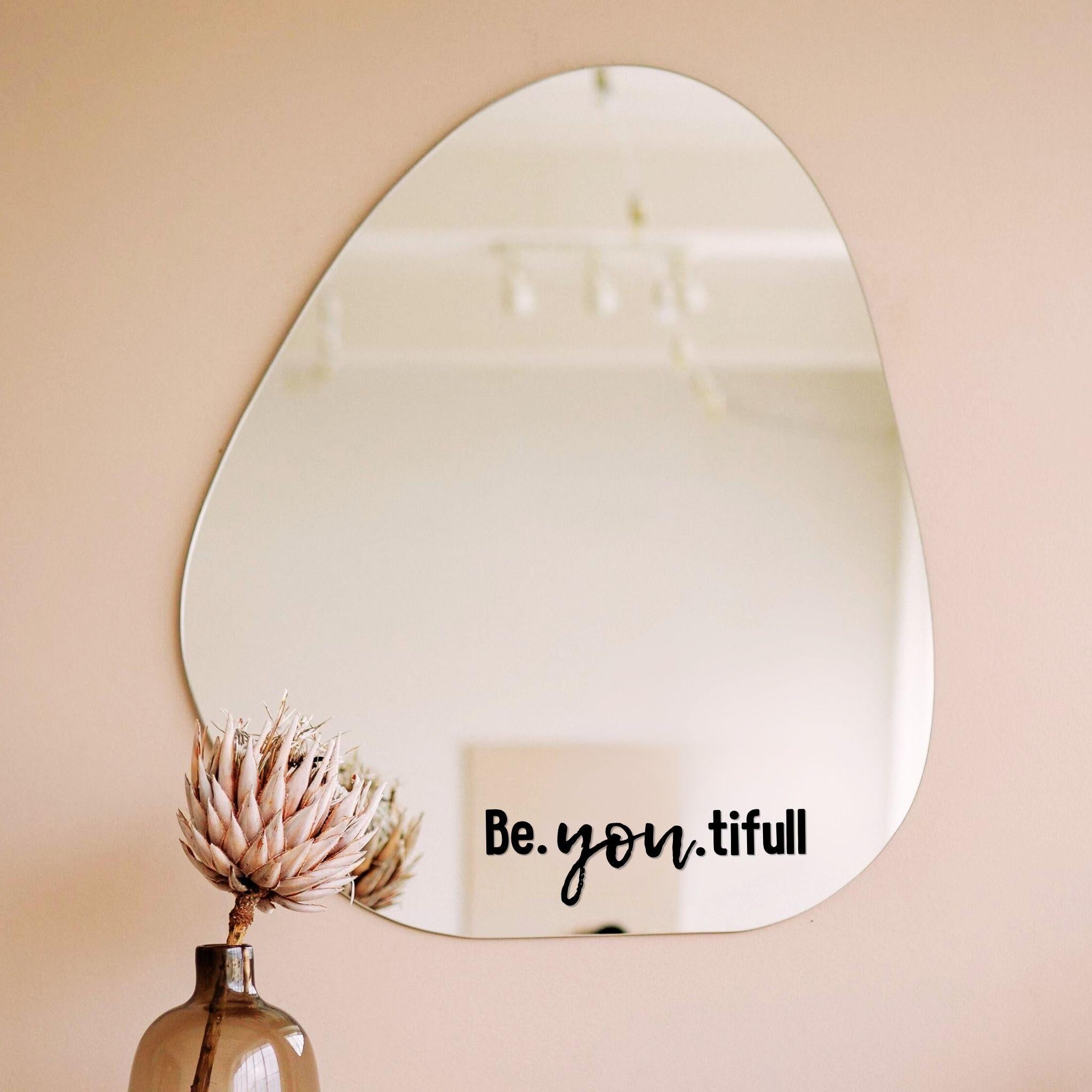 Zwarte spiegelsticker met tekst Be.you.tiful op een organische spiegel in een licht interieur met vaas en droogbloem