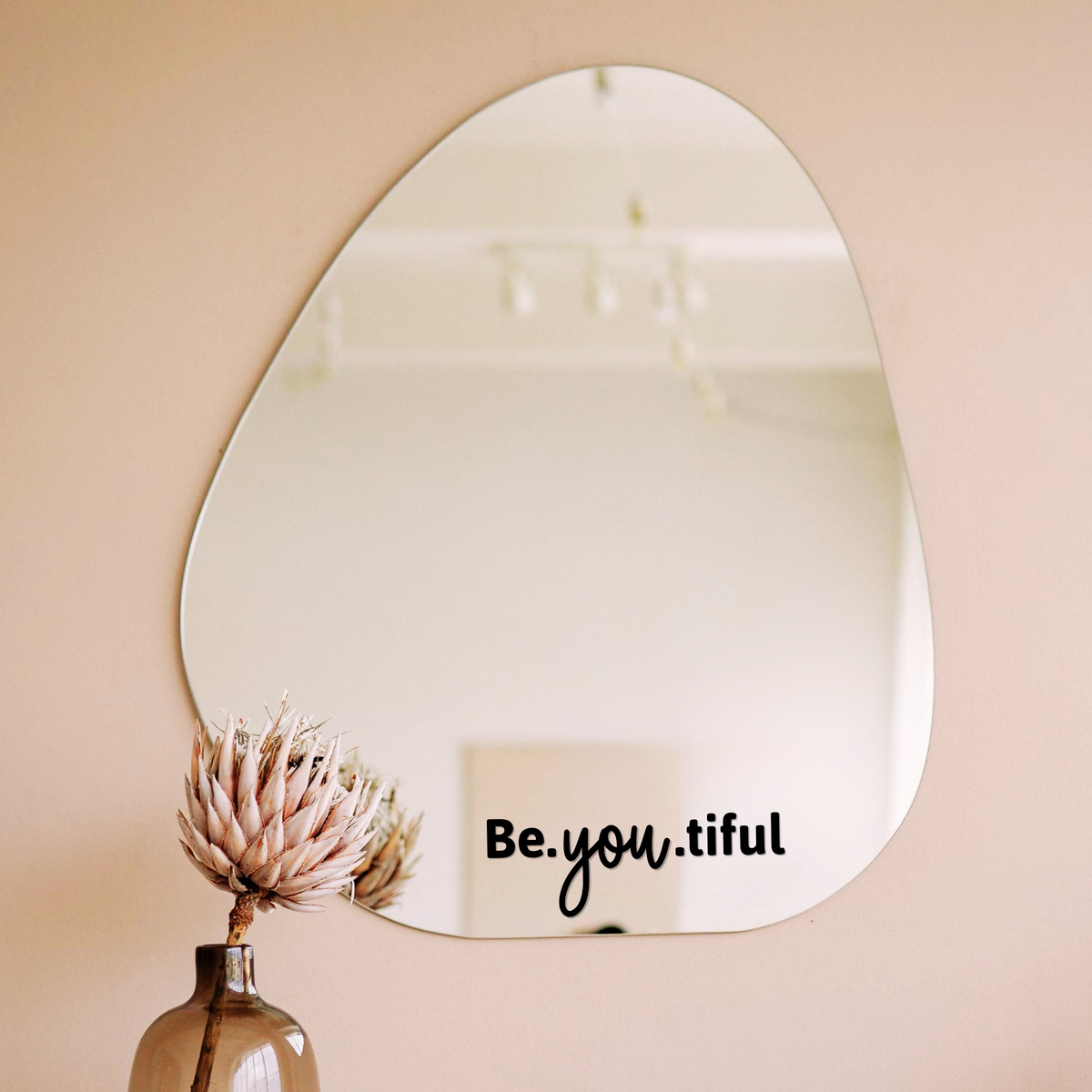 Zwarte spiegelsticker met tekst Be.you.tiful op een organische spiegel in een licht interieur met vaas en droogbloem