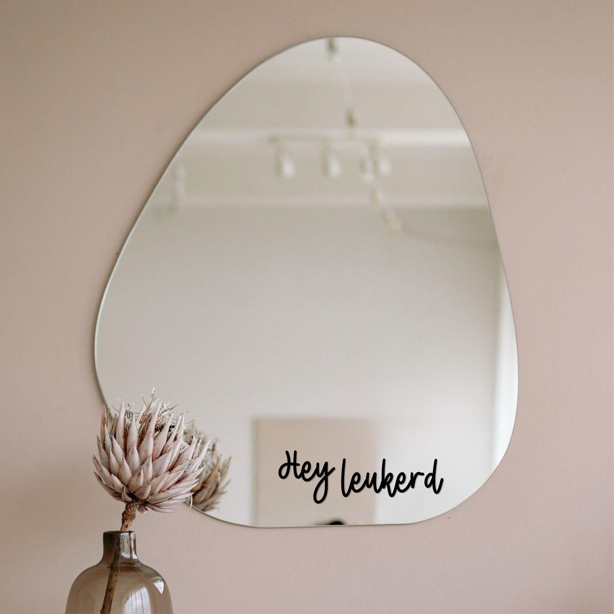 Zwarte spiegelsticker met tekst Hey leukerd op een organische spiegel in een licht interieur met vaas en droogbloem