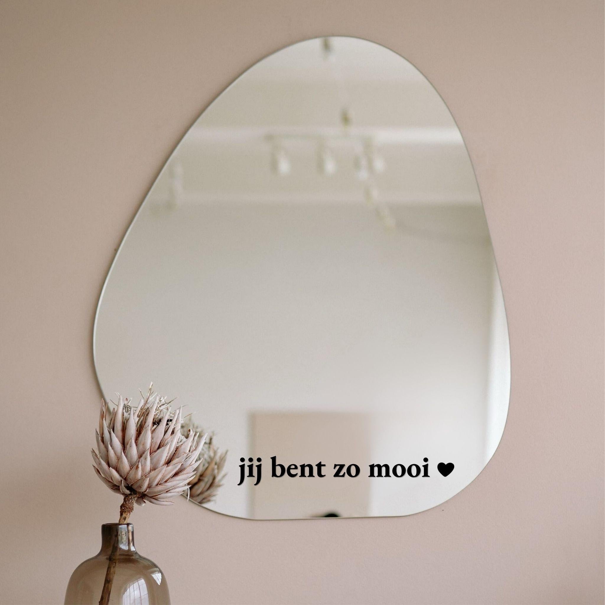 Zwarte spiegelsticker met tekst jij bent zo mooi met hartje op een organische spiegel in een licht interieur met vaas en droogbloem
