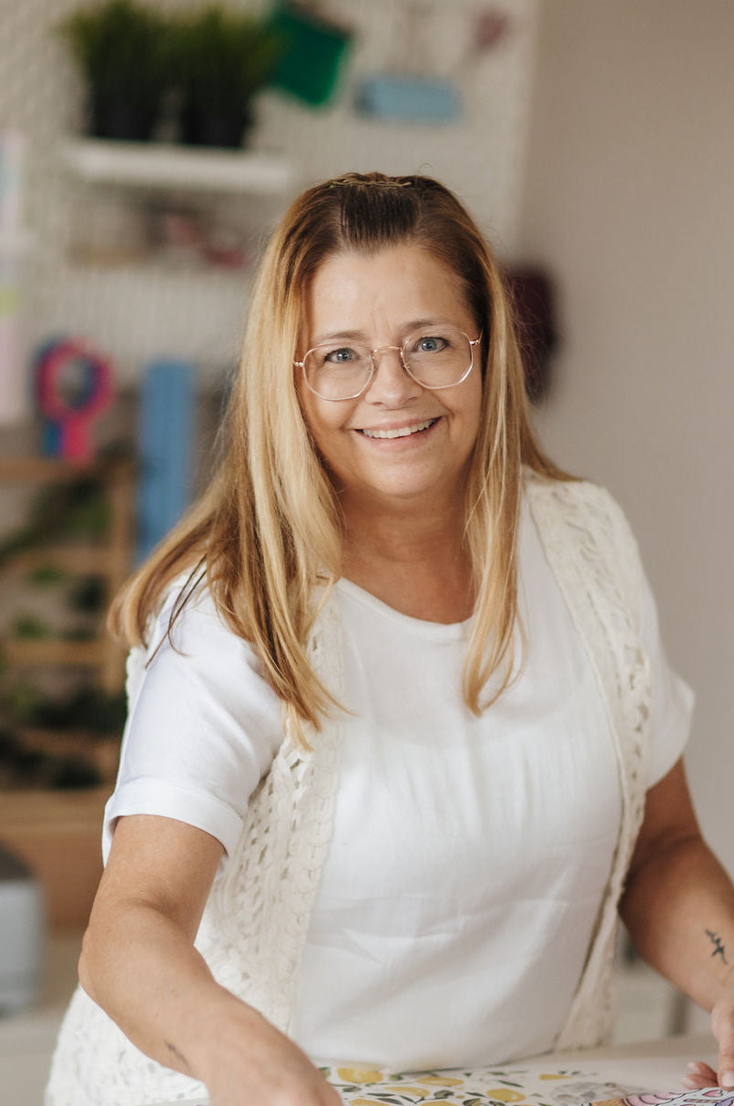 Portret van vrouw in lichte kleding met bril, lachend in een creatieve werkruimte.