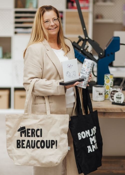 Vrouw toont gepersonaliseerde producten zoals tote bags, mokken, tumblers en een tegel in een creatieve werkruimte.
