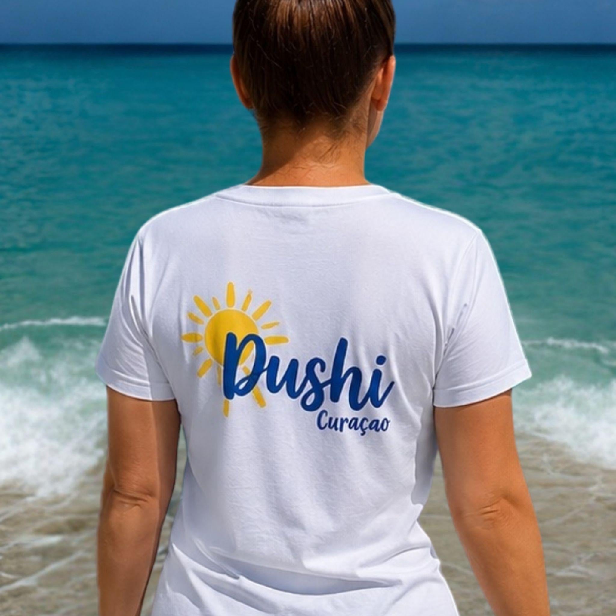 Vrouw draagt een wit Dushi Curaçao T-shirt op het strand met de rugopdruk 'Dushi Curaçao' en een gele zon.